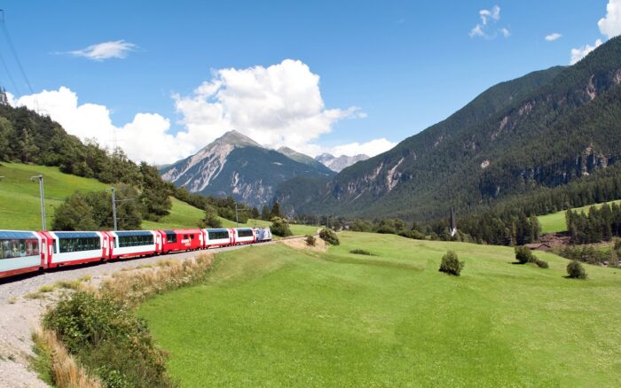 A red passenger train traveling through lush green fields under a wide blue sky
